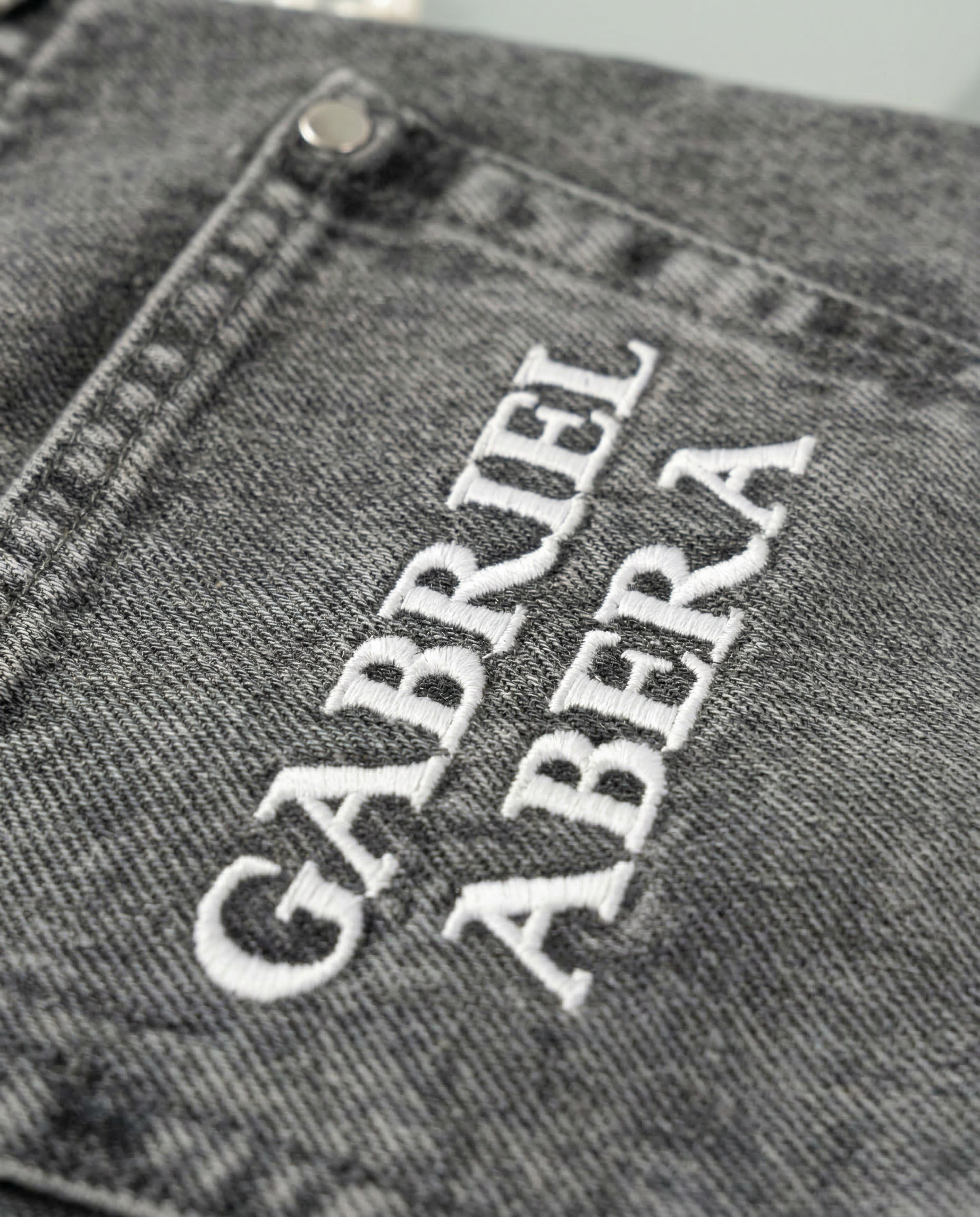 Close-up of embroidered GABRIEL ABERA logo on the back pocket of grey rhinestone denim jeans.