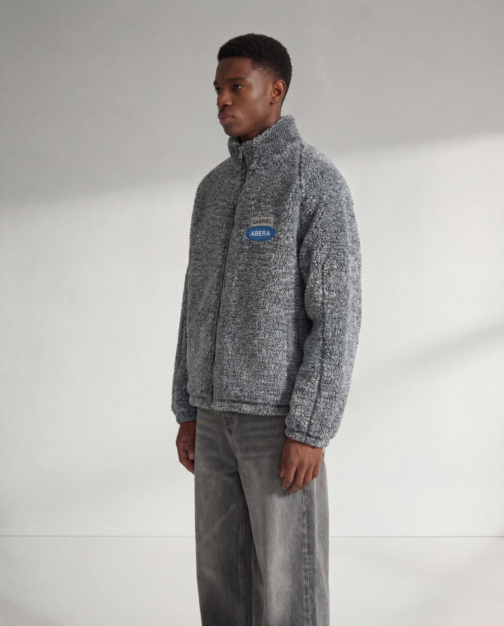 Graue GABRIEL ABERA Fleecejacke auf männlichem Model in Seitenansicht, weiches Fleece-Material, relaxed Streetwear Fit und Logo-Patch auf der Brust sichtbar.