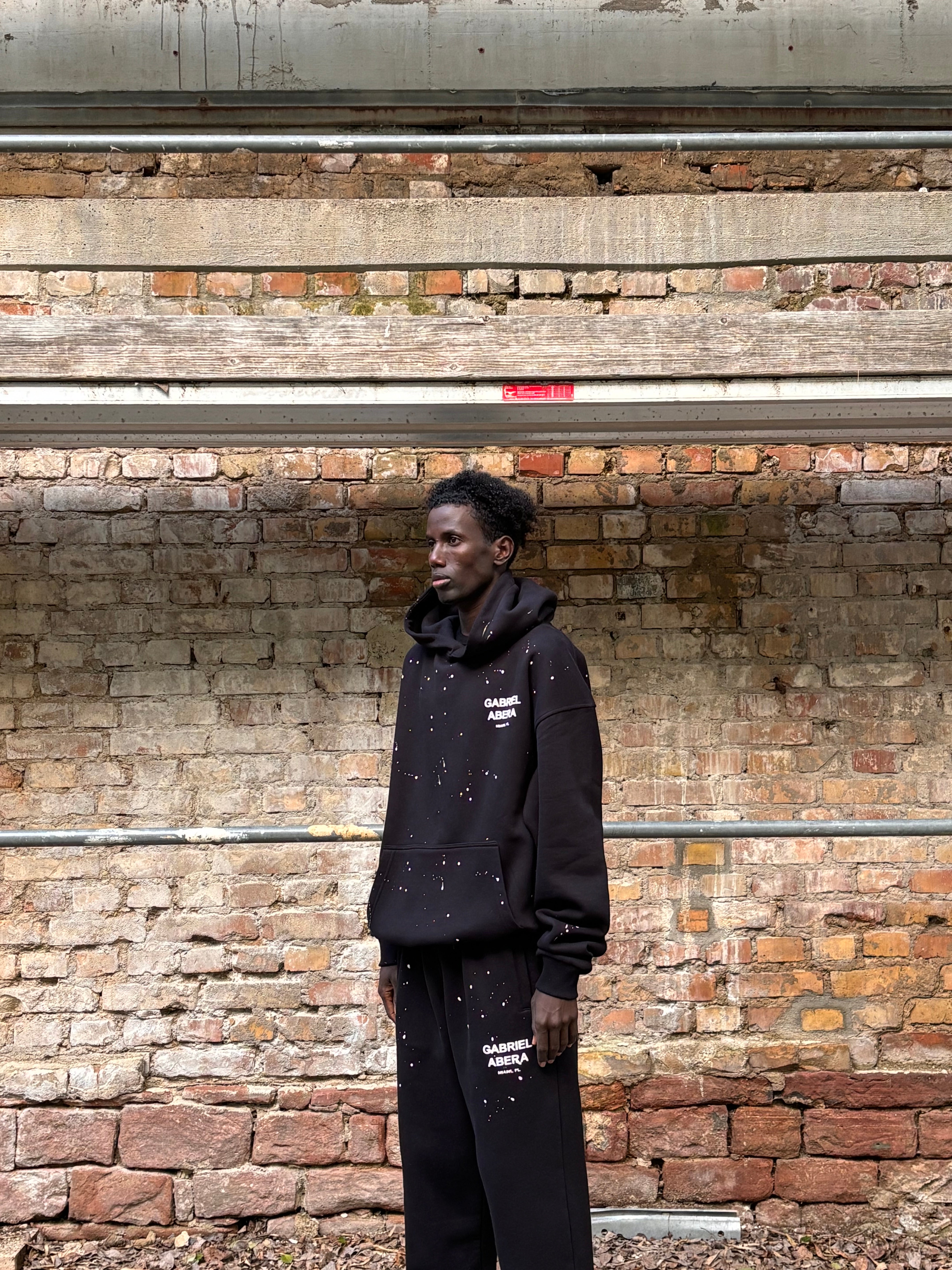 Model trägt GABRIEL ABERA Splattered Puff Hoodie Black/White – Seitenansicht, lockerer Oversized Fit und künstlerisches Paint Splatter Design.