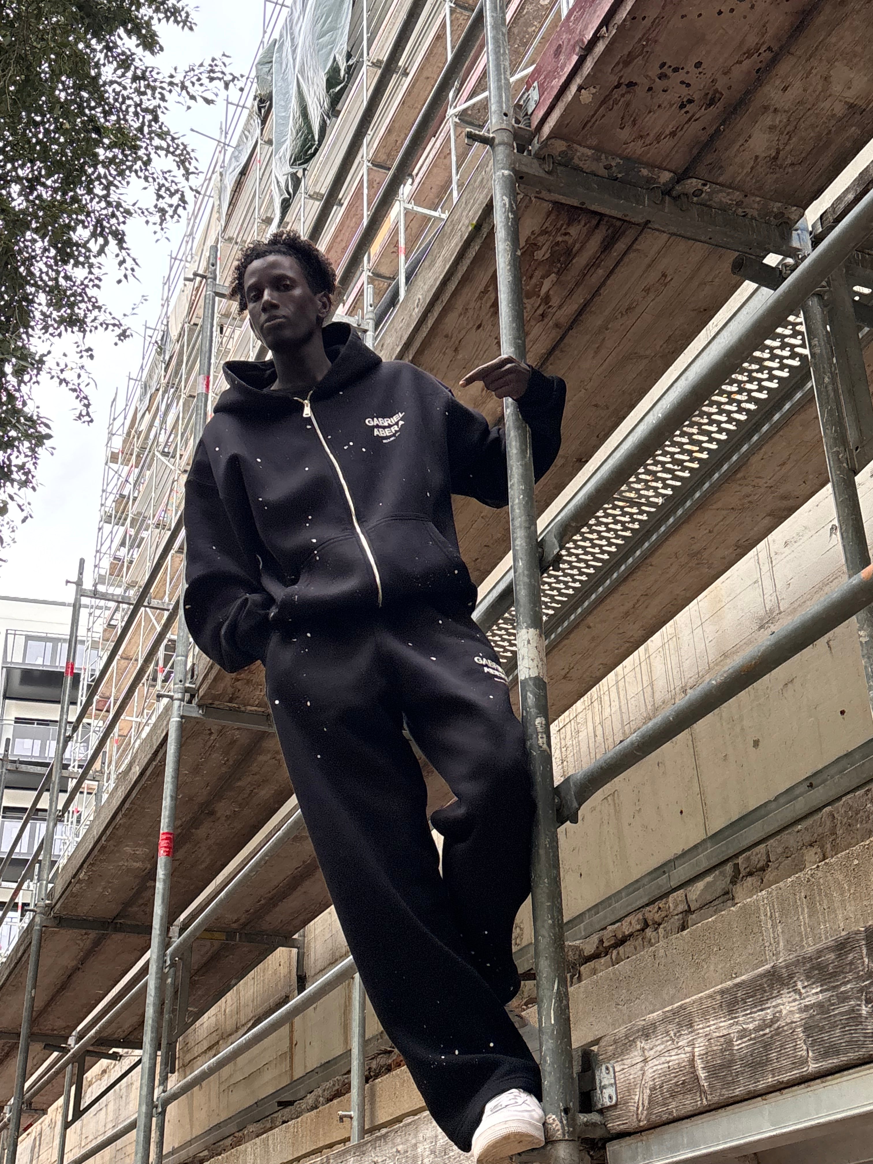 Model trägt GABRIEL ABERA Splattered Puff Zip Hoodie und die passende Jogginghose Black/White als passendes Set.