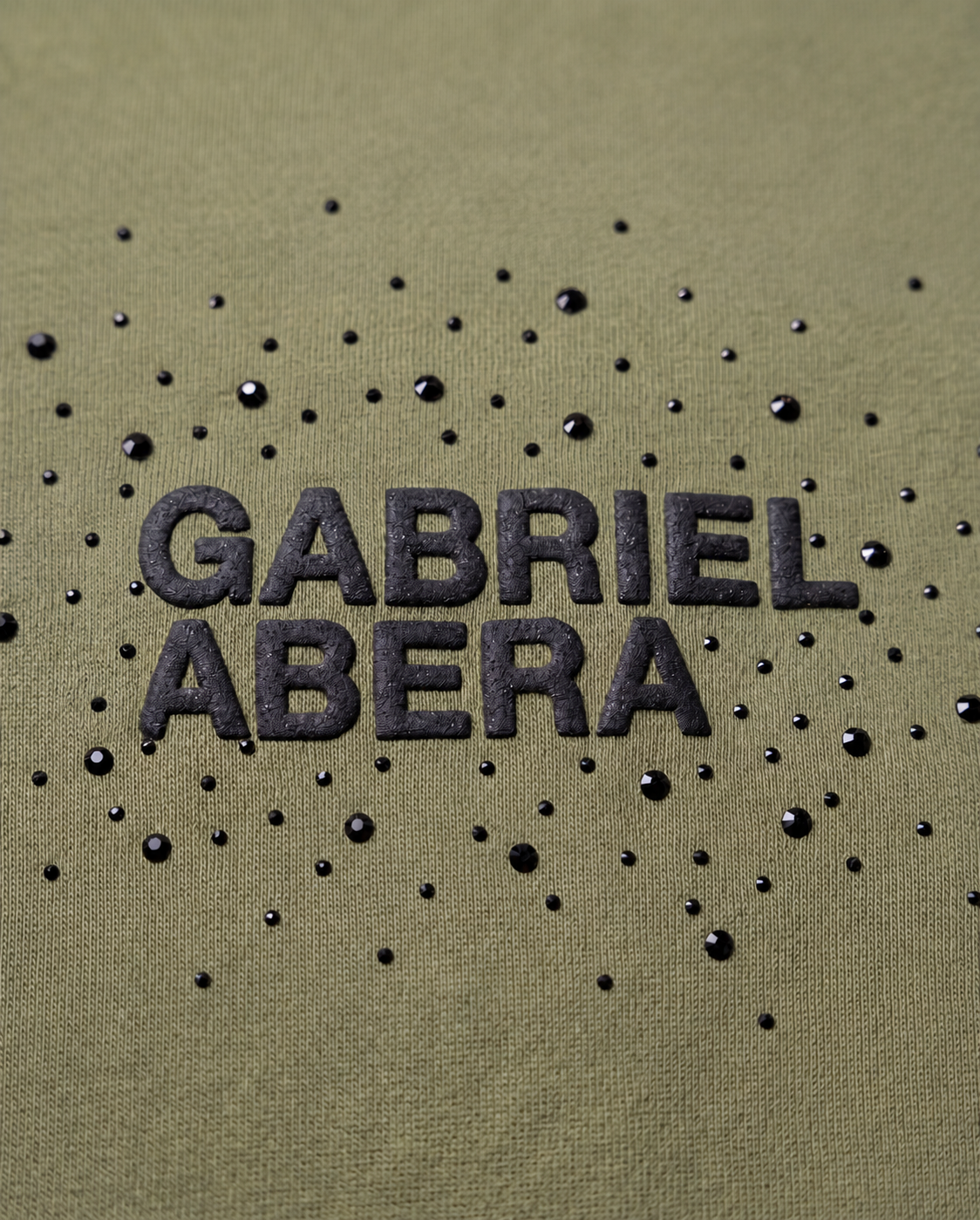 Close-up of the rhinestone 'Gabriel Abera' logo on the olive tee – scattered crystal lettering on washed olive cotton fabric