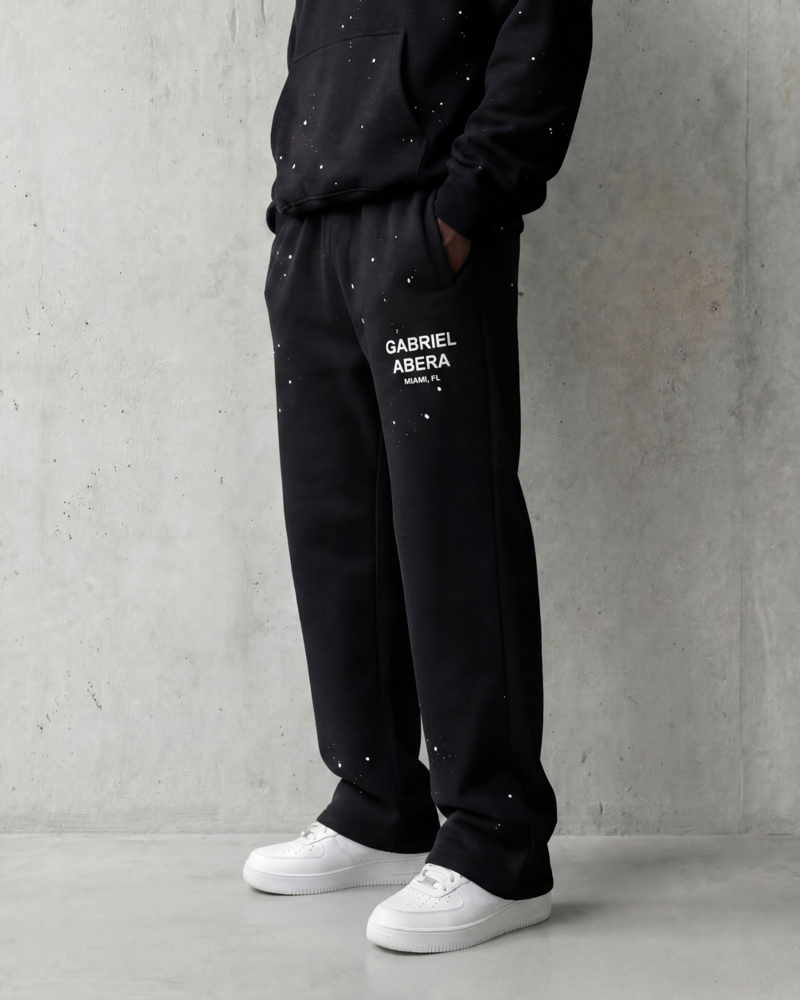 Black oversized sweatpants with paint splatter pattern, straight leg fit, styled with white sneakers, minimal grey background