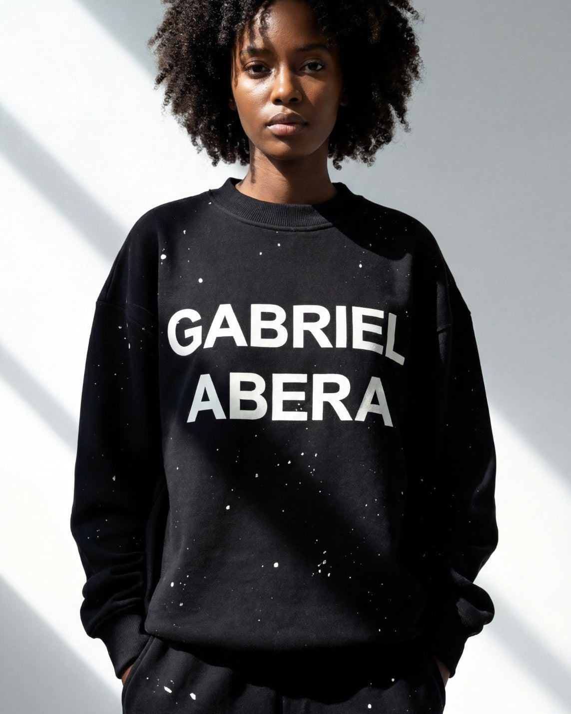 Schwarzes weibliches Model trägt den GABRIEL ABERA Sweater in Schwarz mit handgesprühten weißen Farbspritzern im Oversized Streetwear Fit, Frontansicht.