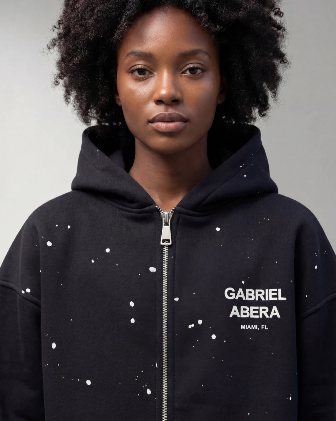 Schwarzes weibliches Model trägt den GABRIEL ABERA Zip Hoodie in Schwarz mit splattered Frontdesign, Streetwear Frontansicht.
