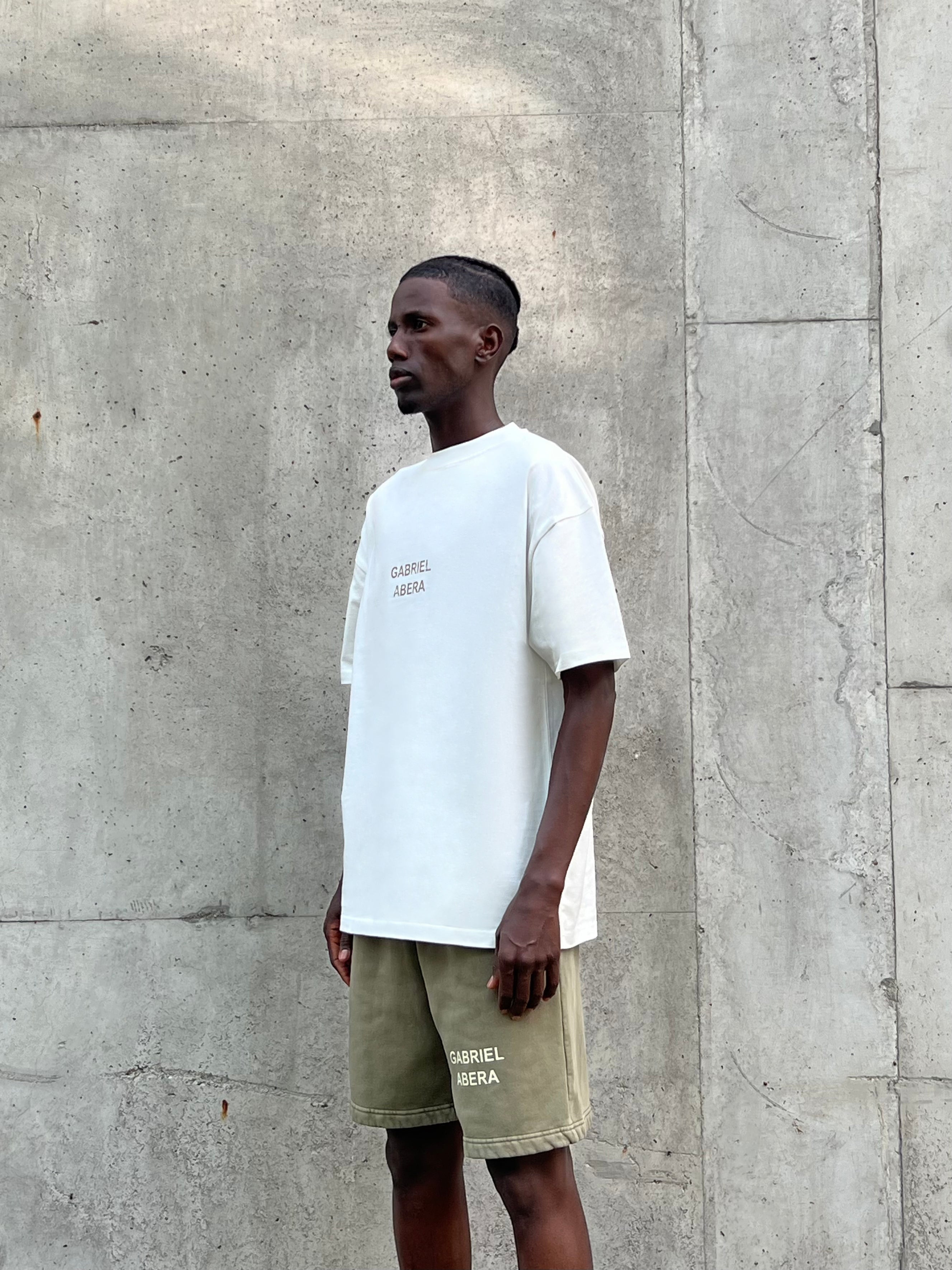 Model wearing cream white GA T-Shirt with khaki GA logo and matching khaki shorts, standing against concrete wall, side-front view