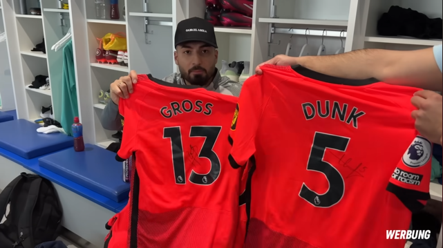 Load video: German Influencer Diyar holds up a signed Pascal Groß Shirt next to a signed Leis Dunk shirt while wearing the Gabriel Abera Cap