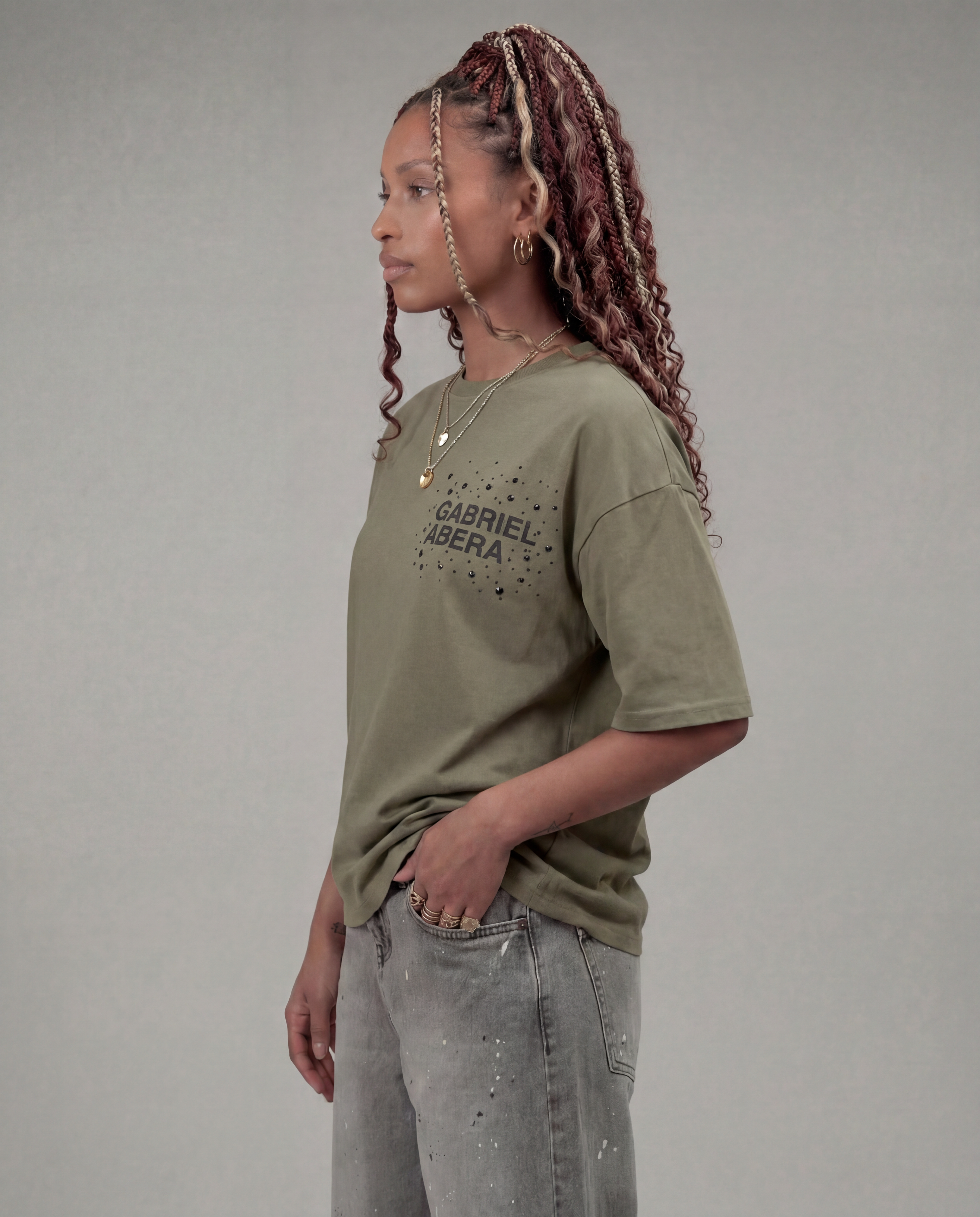 Female model wearing the Gabriel Abera Rhinestone Logo Tee in olive / green with denim shorts – three-quarter side view, relaxed fit, grey background