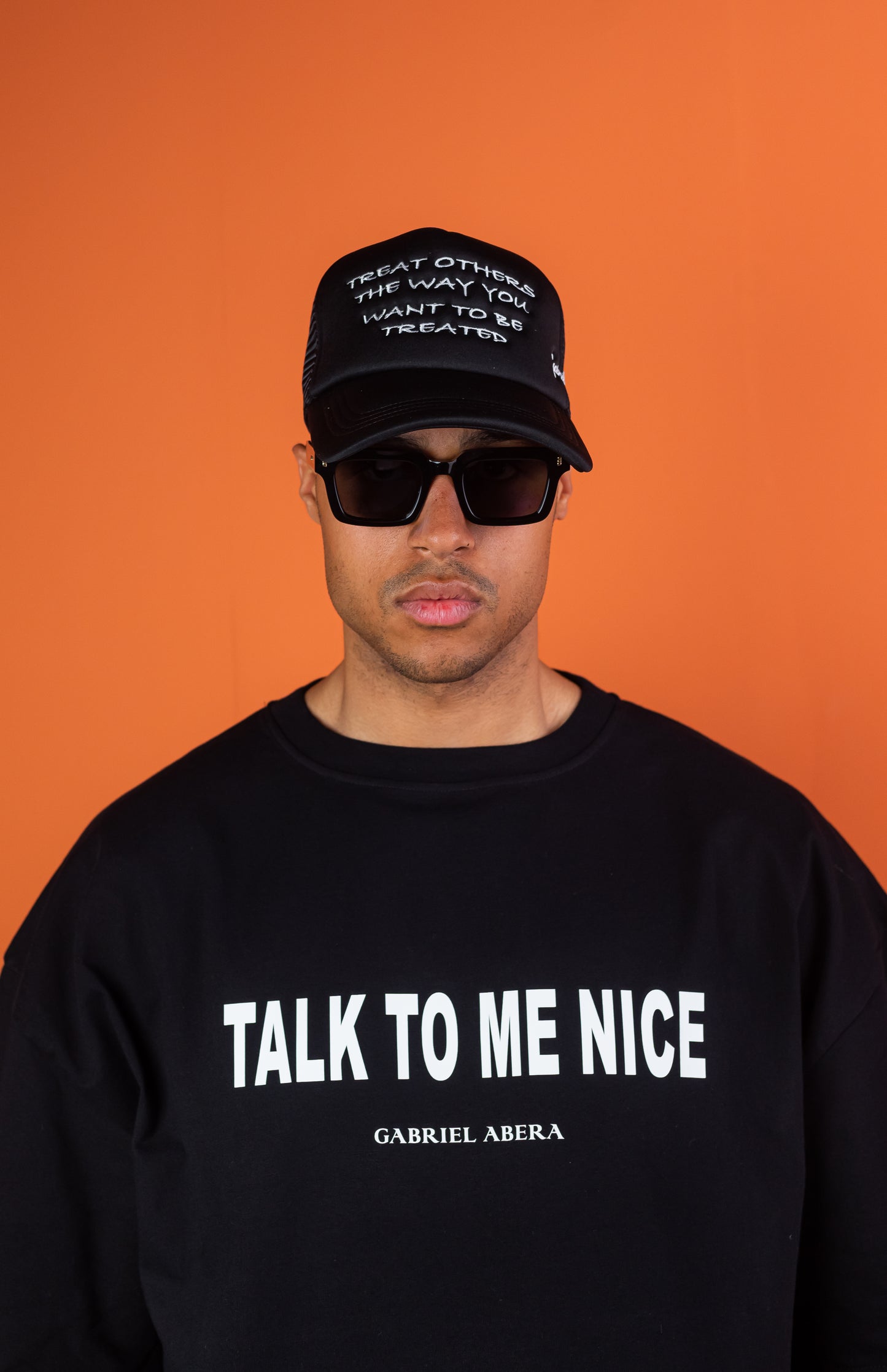 Vorderansicht eines männlichen Models, das ein schwarzes Oversize Tshirt mit der weissen Aufschrift Talk to me Nice und eine schwarze Kappe mit weisser Treat others the way you want to be threated Aufschrift trägt.