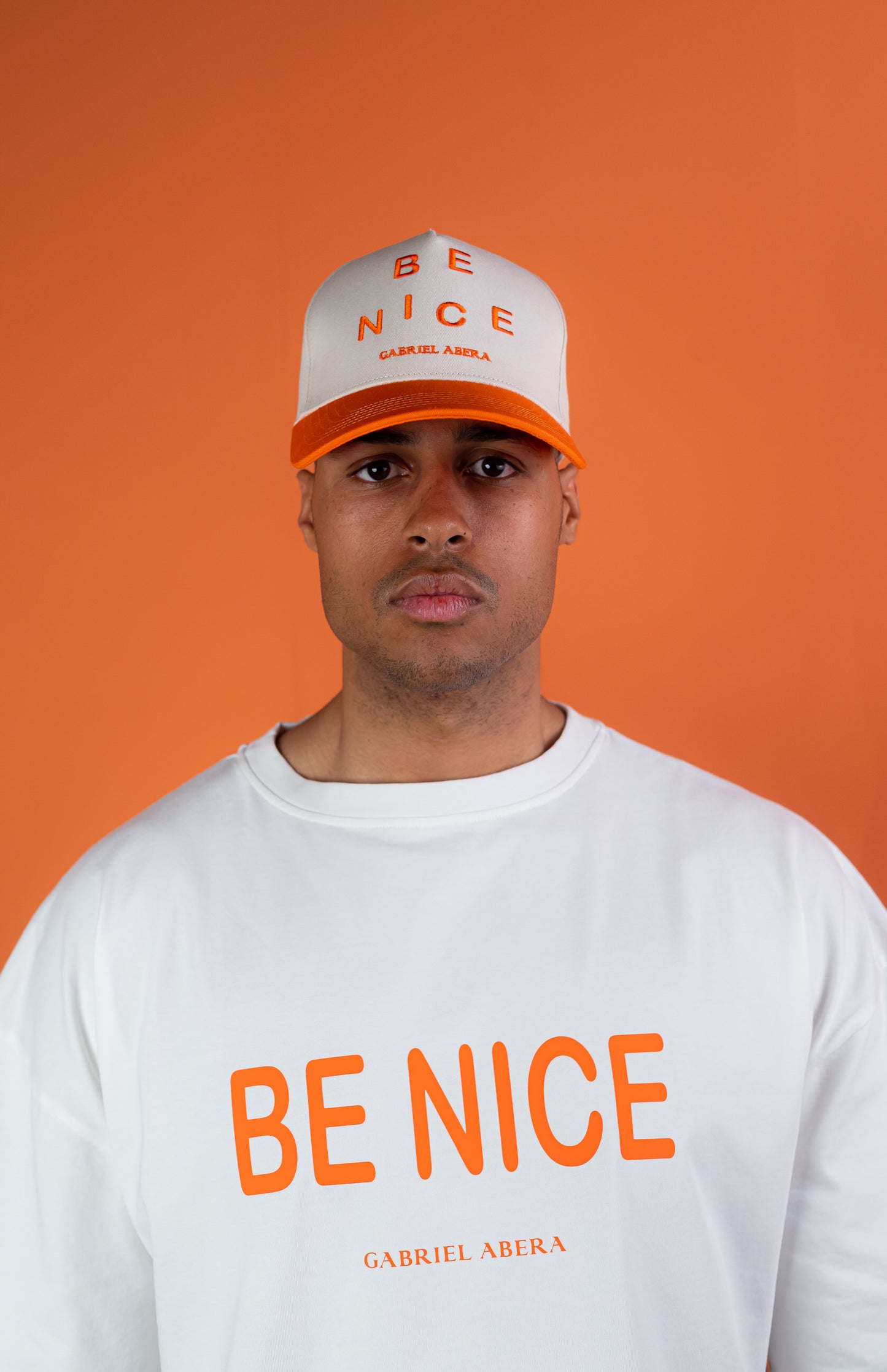Male model wearing a White cap with orange shield with "be nice" writing on the front