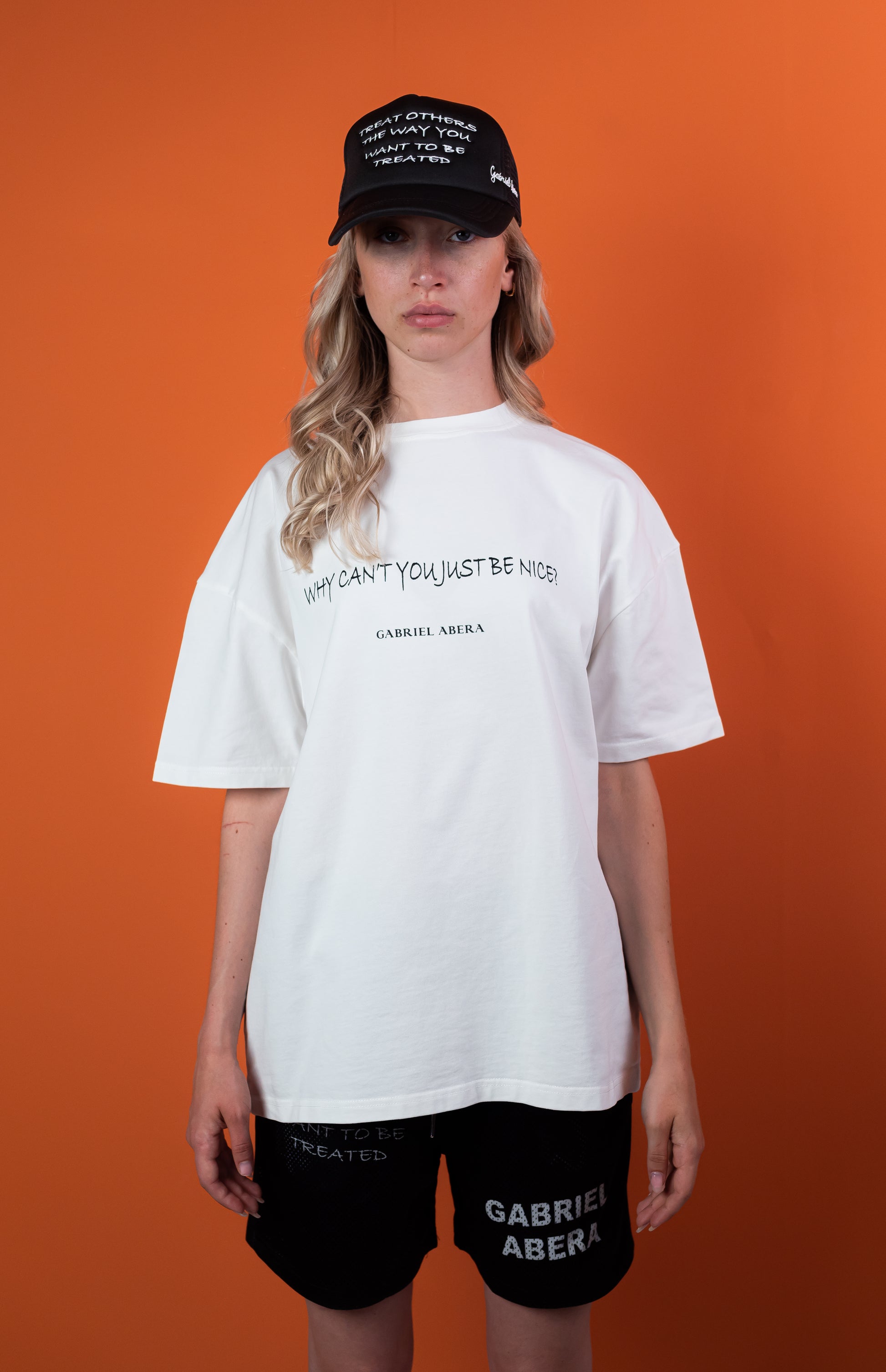 Vorderansicht eines weiblichen Models, das ein weißes Oversize Tshirt mit der schwarzen Aufschrift Why can't you just be nice, eine schwarze Mesh shorts mit einem weissen Gabriel Abera Logodruck und eine schwarze Kappe mit weisser Treat others the way you want to be threated Aufschrift trägt.