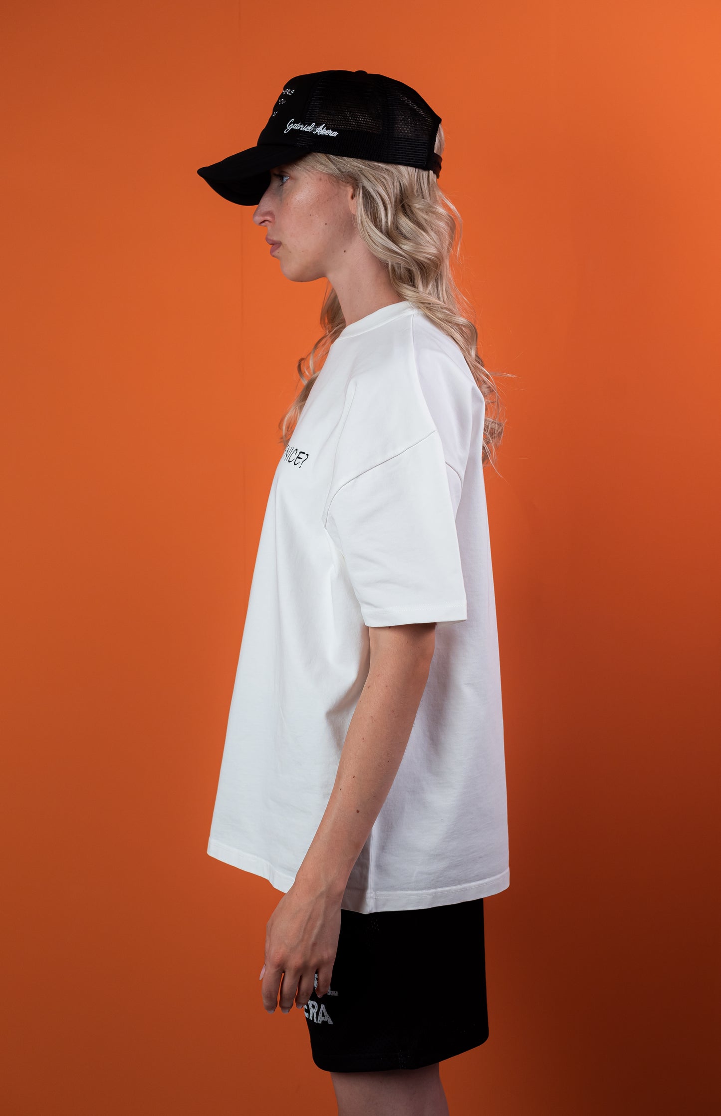 Seitenansicht eines weiblichen Models, das ein weißes Oversize Tshirt mit der schwarzen Aufschrift Why can't you just be nice, eine schwarze Mesh shorts mit einem weissen Gabriel Abera Logodruck und eine schwarze Kappe mit weisser Treat others the way you want to be threated Aufschrift trägt.