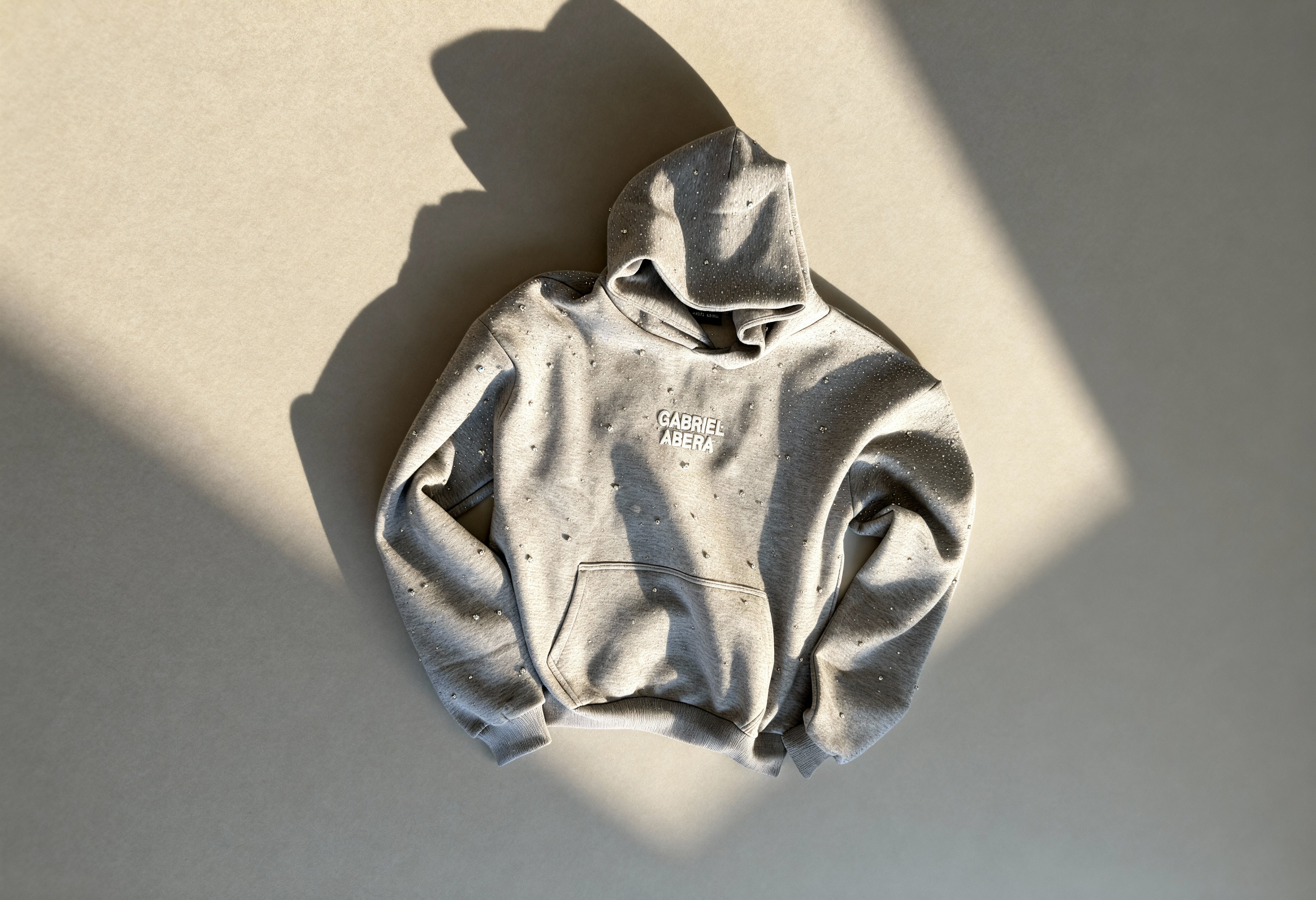 GABRIEL ABERA Hoodie in grau mit Rhinestones auf dem Boden liegend.