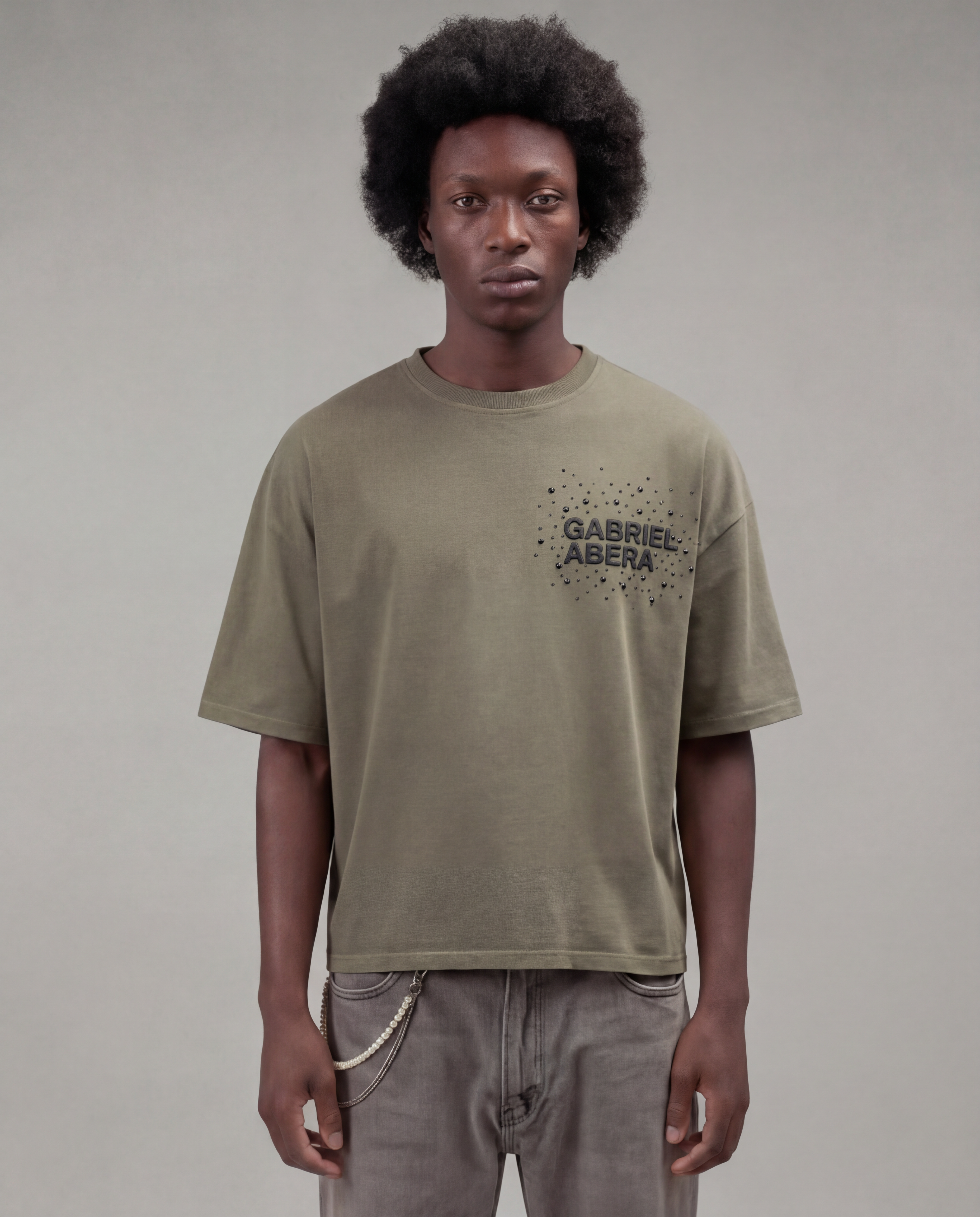 Male model with afro wearing the Gabriel Abera Rhinestone Logo Tee in olive / green – oversized boxy fit, front view, black rhinestones 'Gabriel Abera' logo on chest, grey background