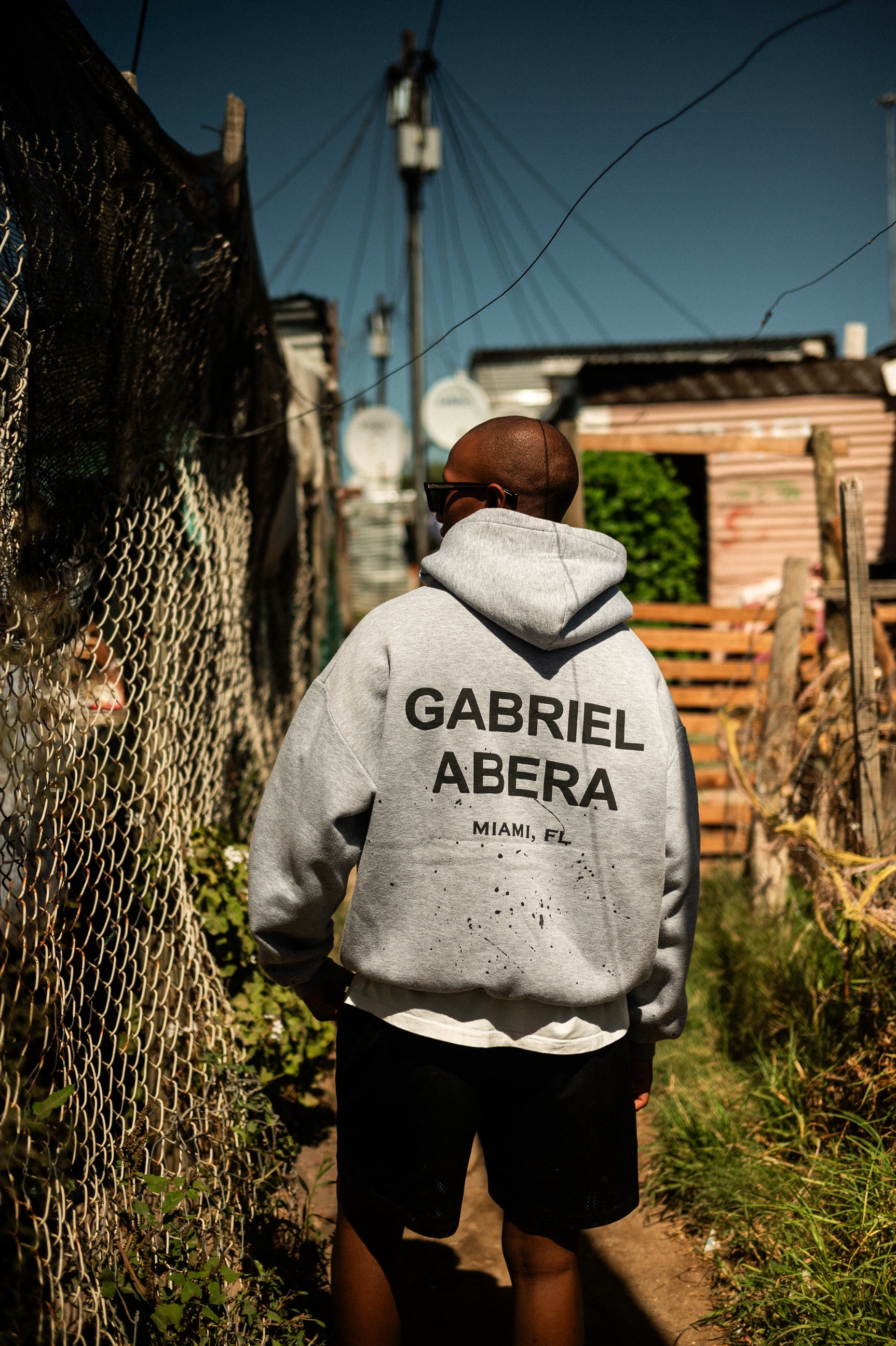 Model in GABRIEL ABERA Hoodie vor Wellblechhütte in Lwandle.