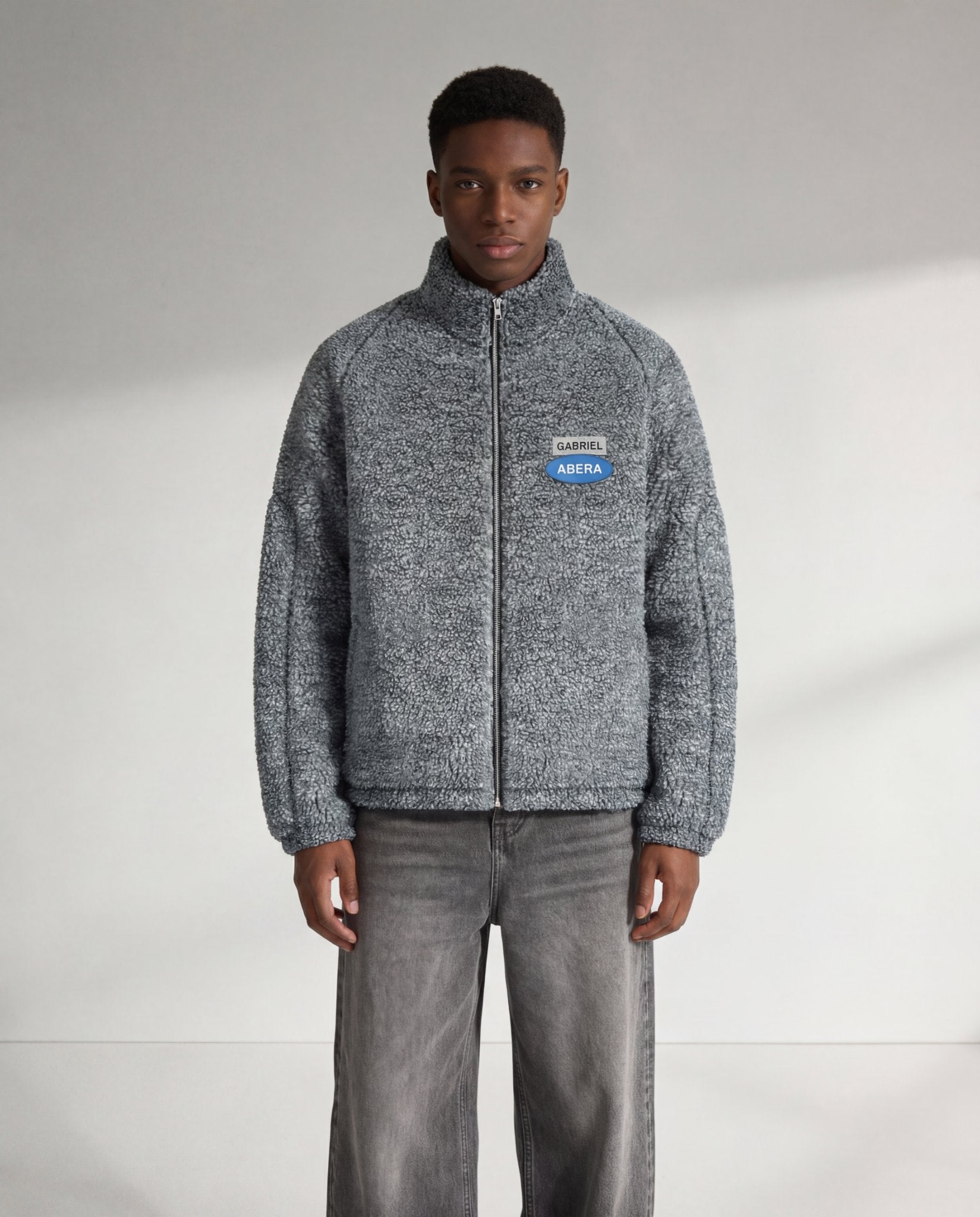 Graue GABRIEL ABERA Fleecejacke auf männlichem Model von vorne, Frontansicht mit Reißverschluss, Logo-Patch und lockerer Passform.