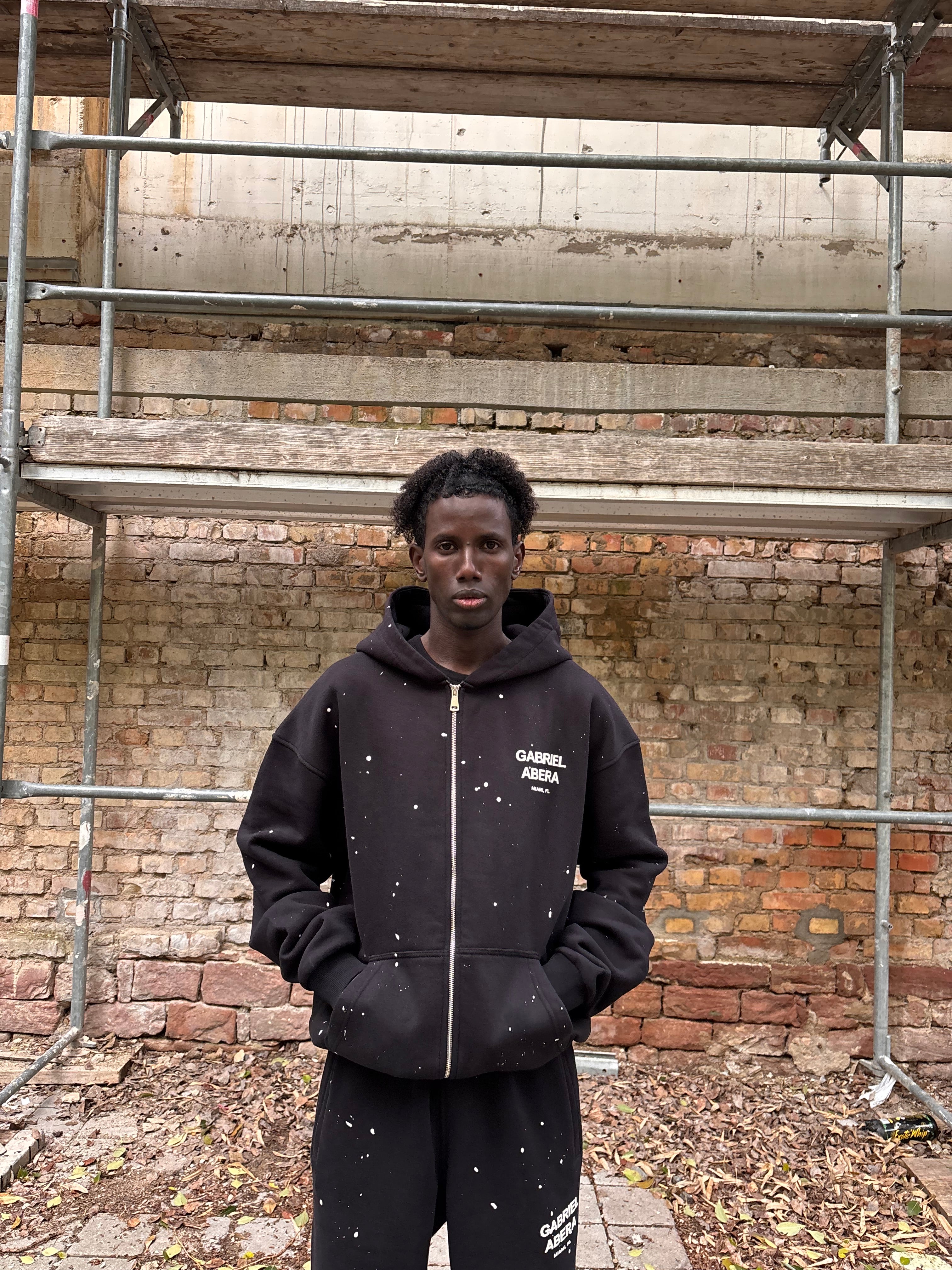 Model trägt GABRIEL ABERA Splattered Puff Zip Hoodie Black/White – Frontansicht, Reißverschluss offen/loss.