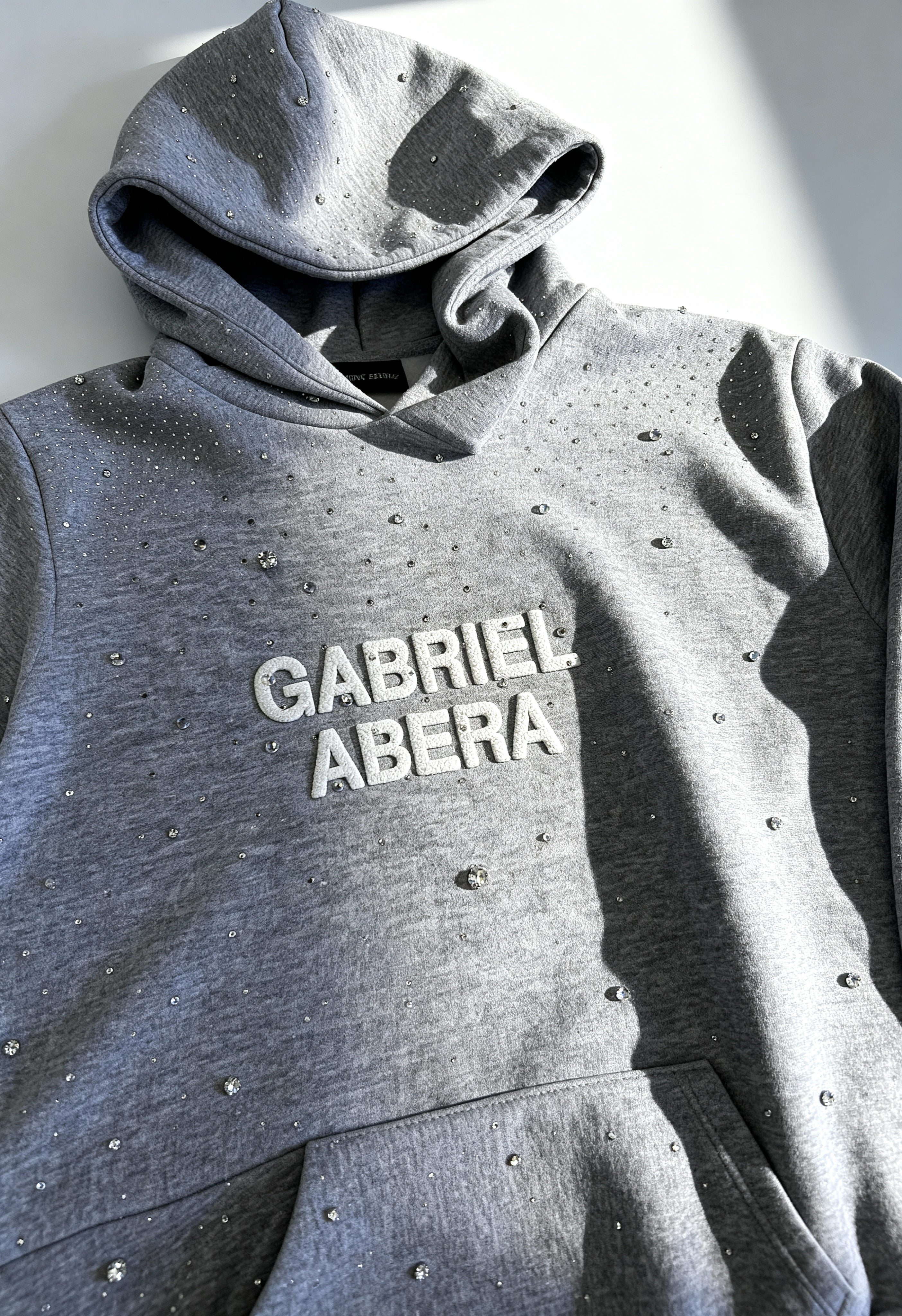 Nahaufnahme des grauen GABRIEL ABERA Rhinestone Hoodie – handgesetzte Steine funkeln, hochwertige Textur, Fashion Detailshot.