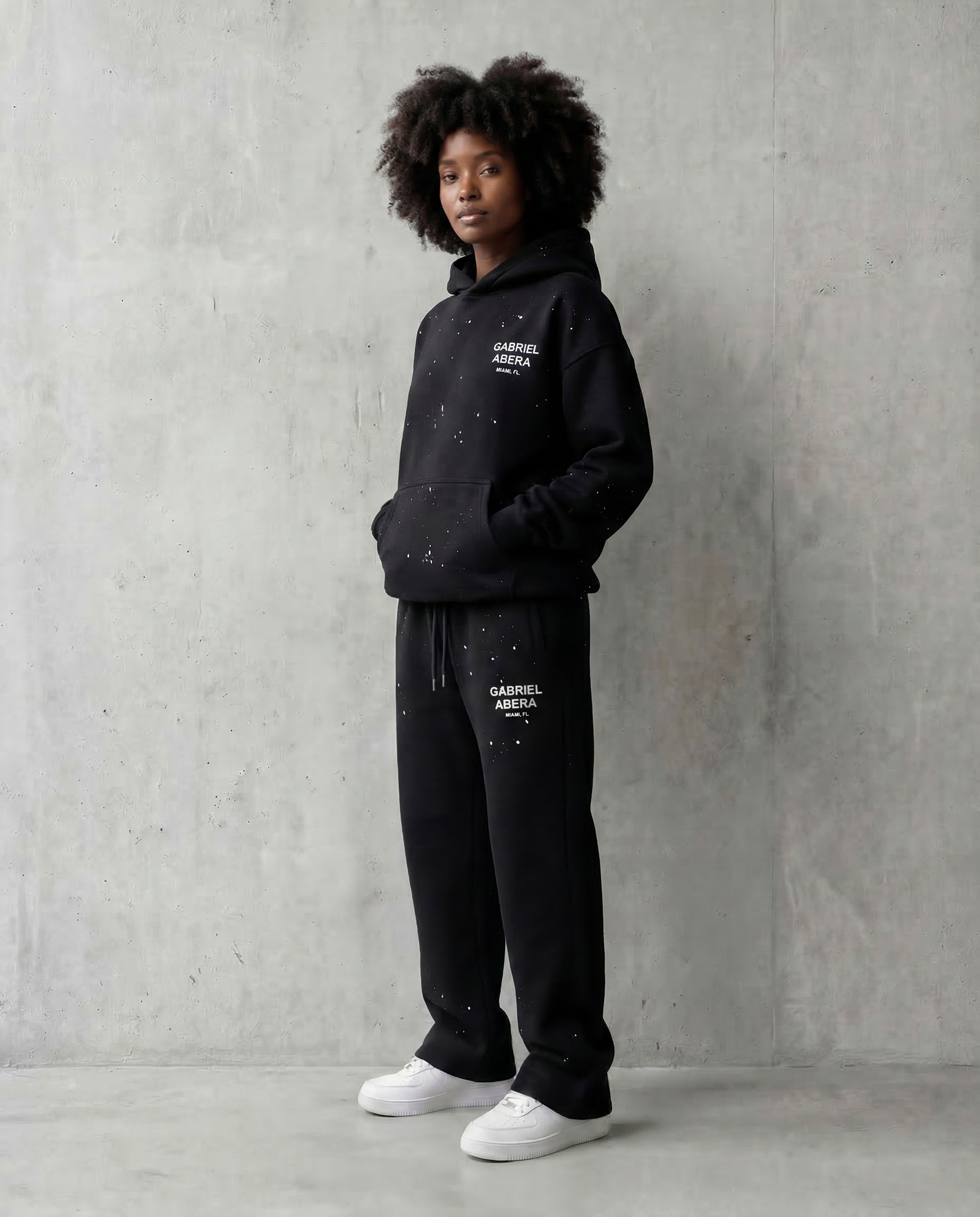 Black paint splatter tracksuit worn by female model, oversized hoodie and straight fit sweatpants, GABRIEL ABERA logo on chest and leg, minimal studio background