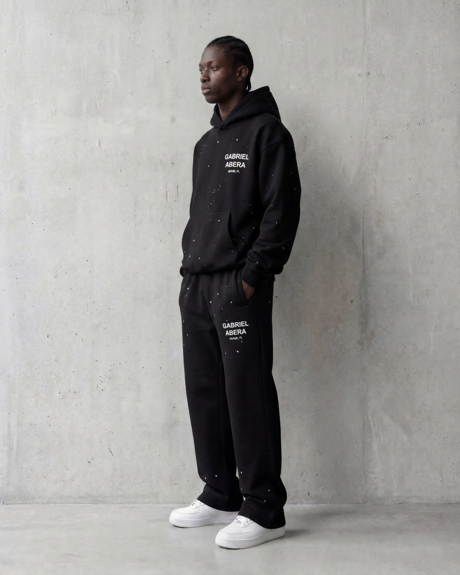 Male model wearing black paint splatter hoodie and sweatpants set by GABRIEL ABERA premium streetwear, side view, relaxed oversized fit, clean studio background