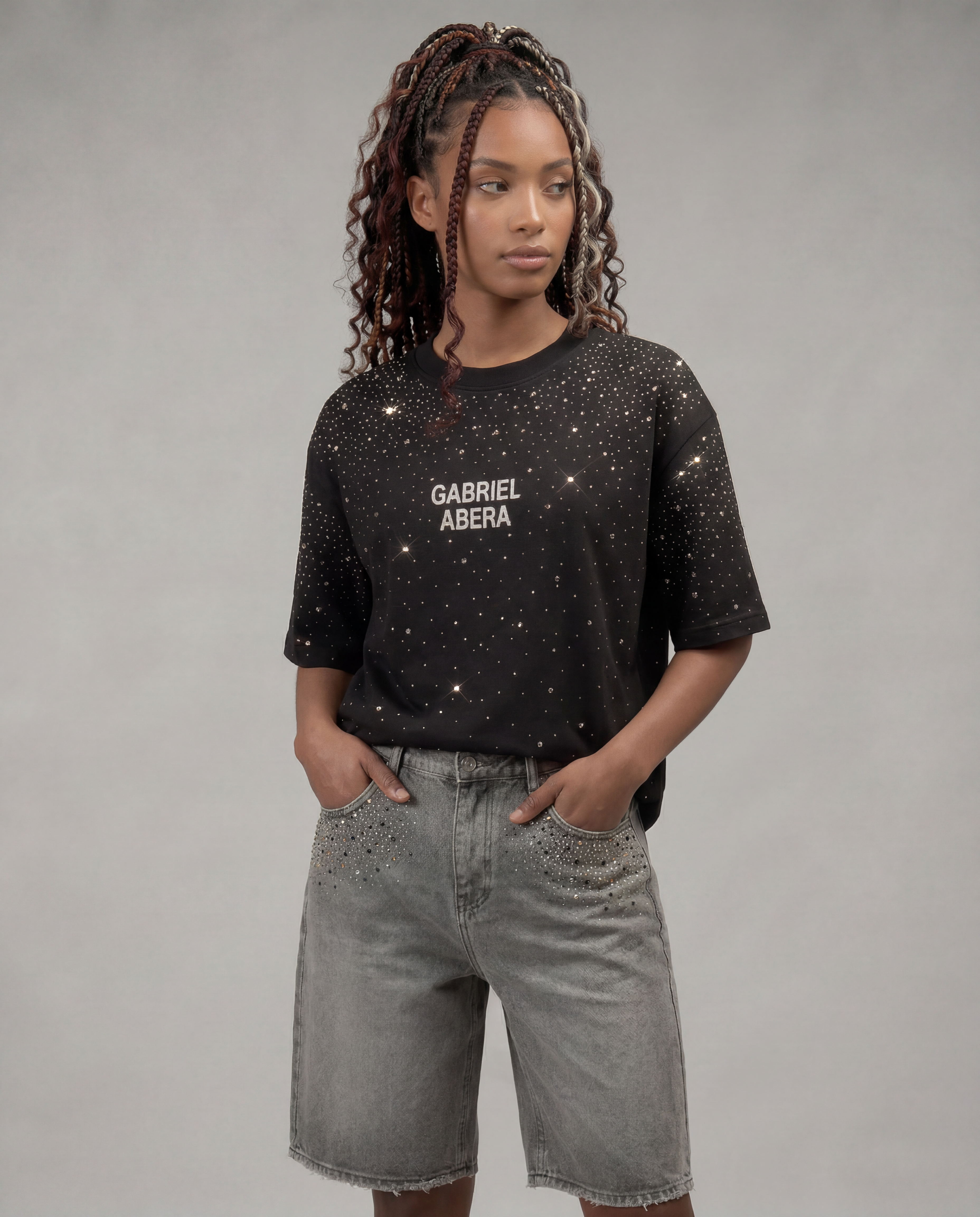 Black oversized "Rhinestone Universe" T-shirt by Gabriel Abera, embellished with hundreds of sparkling rhinestones in varying sizes creating a starry sky pattern. White brand lettering on the chest. Styled with grey denim shorts featuring matching rhinestone detailing. Worn by a female model with braided hair against a grey background.