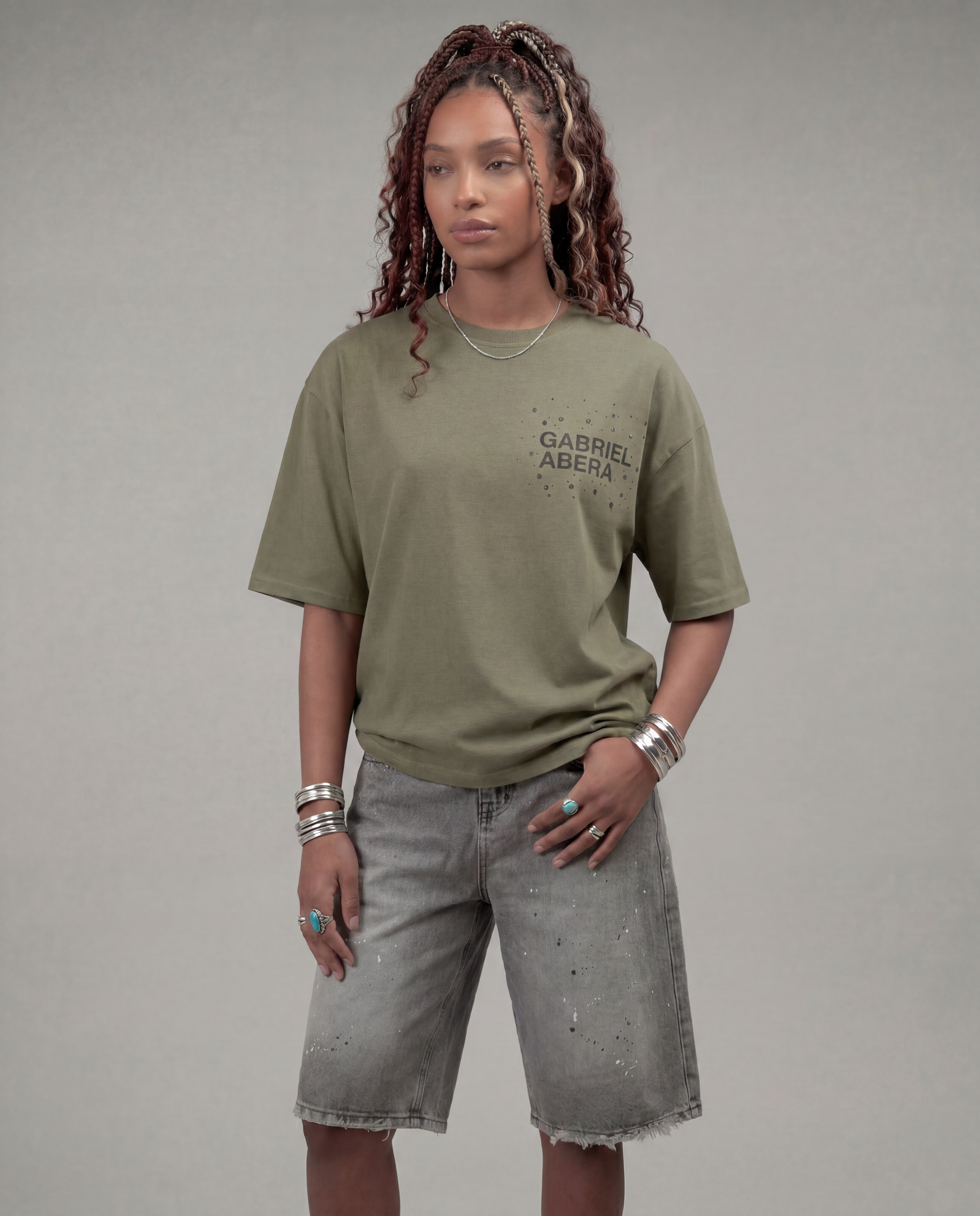 Female model wearing the Gabriel Abera Rhinestone Logo Tee in olive with denim shorts – three-quarter side view, relaxed fit, grey background